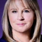 Debbie Foster, a woman wearing a blue shirt and silver necklace with dark blonde shoulder-length hair and a fringe looks at the camera and smiles. The background is dark purple.
