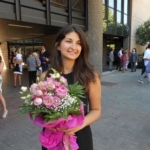 Krizia Ceccobao, a woman in a black dress with dark hair holding pink flowers outside a building.
