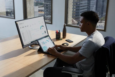 Adult male in an office setting sitting at a desk with his hand poised over the keyboard of a black Microsoft Surface Pro 7 in laptop mode. Microsoft Excel visible on labptop and Microsoft PowerBI screen seen on monitor. Microsoft Surface Arc Mouse shown next to Surface Pro 7.