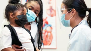 A doctor in face masks and googles talking to a child and her mom, both also in face masks