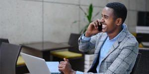 Homem negro sorrindo ao falar no celular com o computador à sua frente