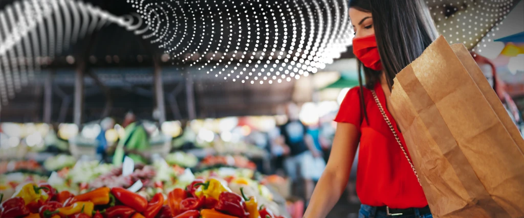 woman in mask at the produce section of a grocery store. She is holding a paper bag and reaching for a red bell pepper