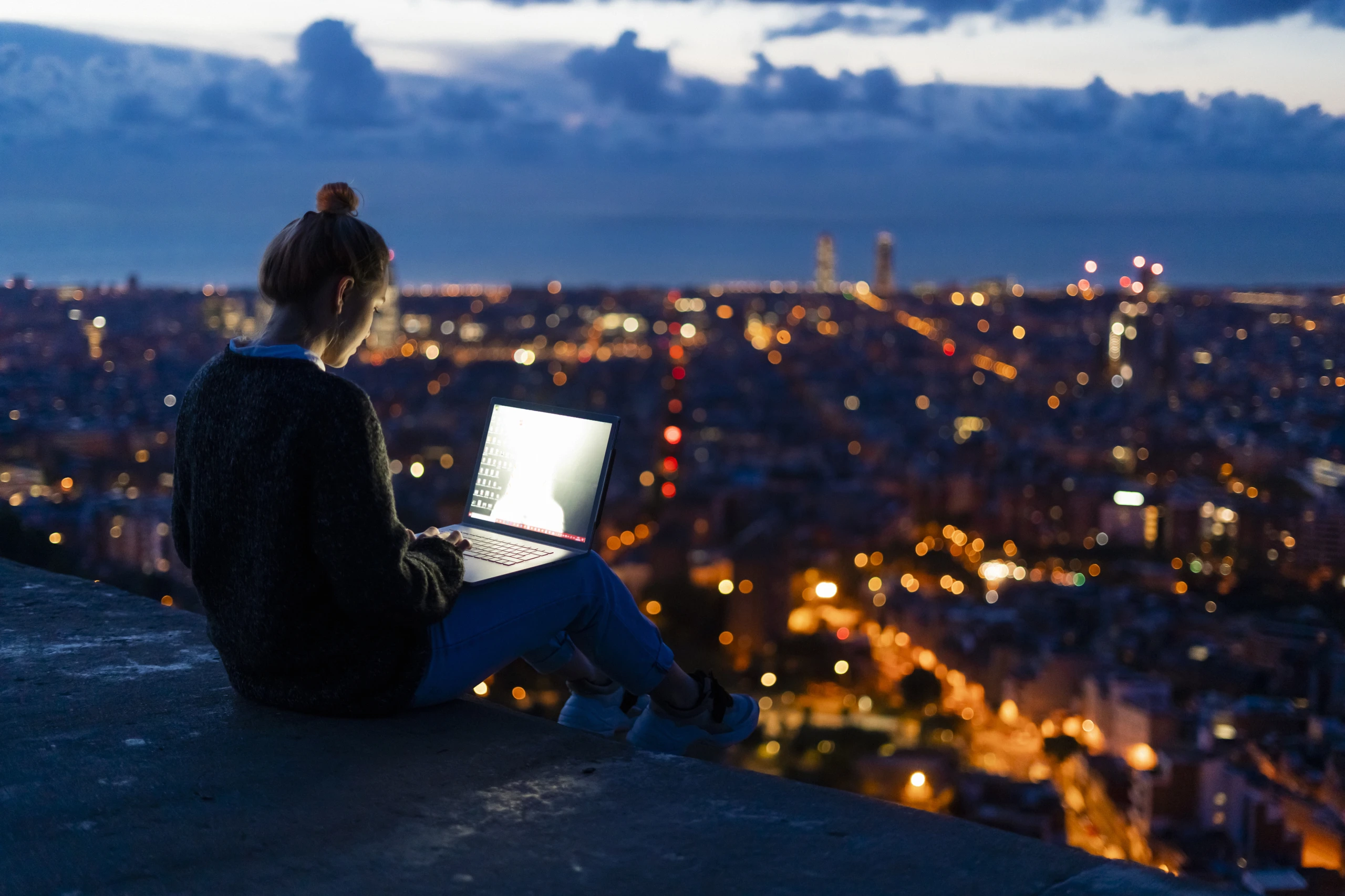 Young woman using laptop at dawn above the city, Barcelona, Spain