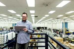 Male factory worker standing above manufacturing plant floor, smiling and looking down at Surface Pro (screen not shown).