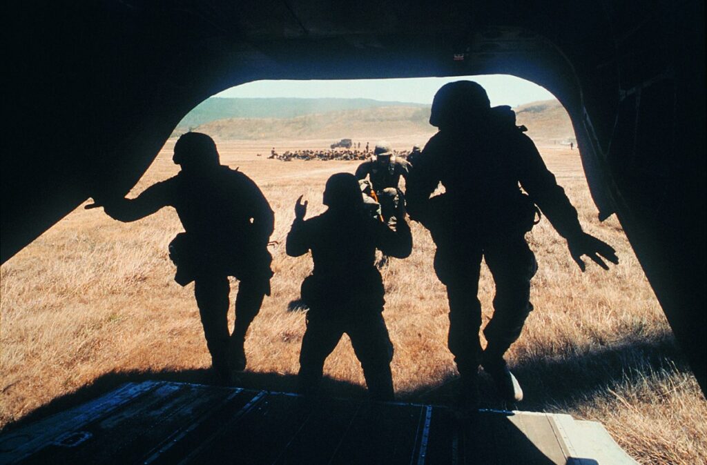Image of defense personnel boarding the back of plane in a remote area, with troops and vehicles in the background. 
