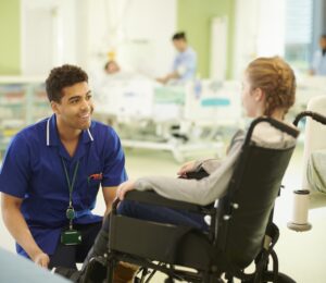 Male paediatric nurse kneeling next to a child in a wheelchair.