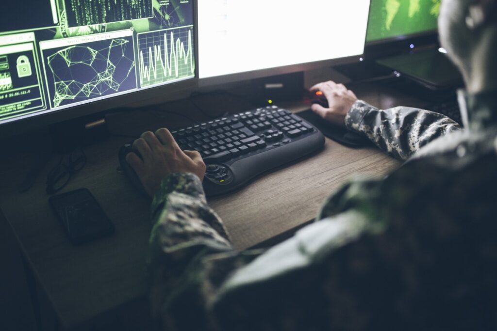 Caucasian male in uniform, sitting at desk with left hand on the keyboard and the right hand on mouse, looking at computer images on a large screen.