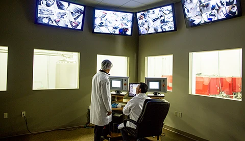 Zwei Männer in einer Art Labor mit einigen Bildschirmen und Schaufenstern. 