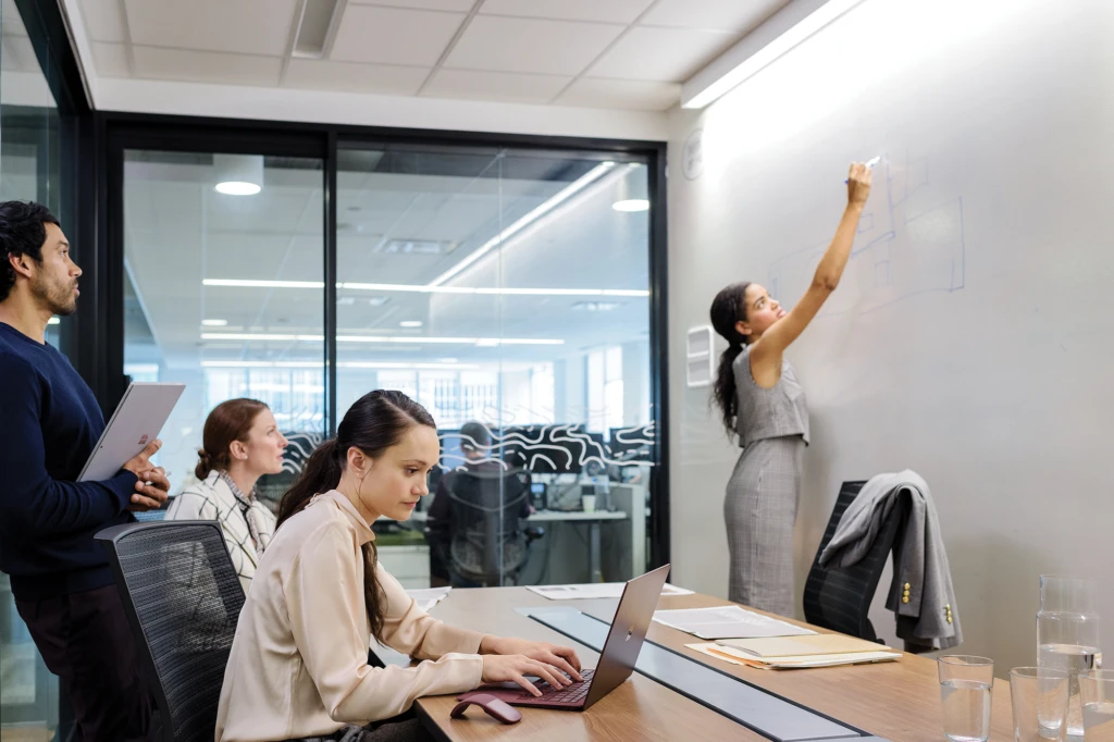 One male and three female coworkers in office conference room. One woman is standing and writing on whiteboard wall. Two other women are seated at table, one using a Surface Laptop (screen not visible) and the other looking at the whiteboard.