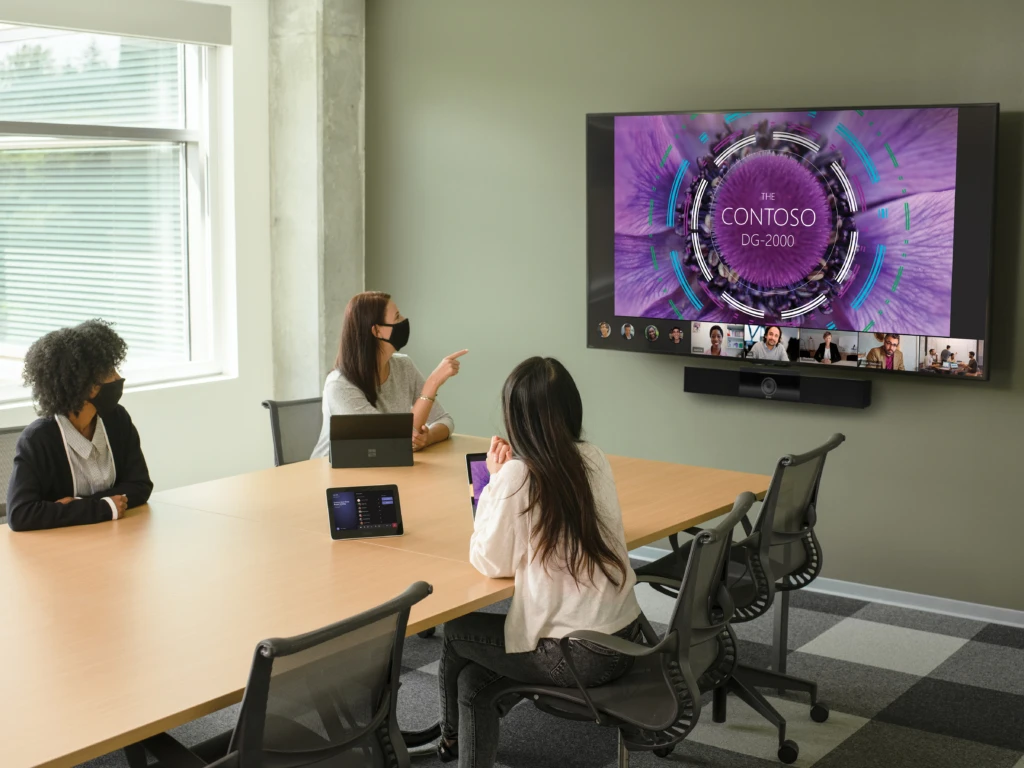 Three females with two wearing masks in a large conference room featuring an Poly Teams Meeting Rooms touch display joined to a Teams Meeting. Screensharing and remote participants shown on a large mounted display and two Surface Devices in view.