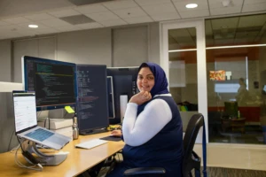 Female developer smiling at camera, wearing a hijab at her desk. Employee experiences are designed to empower everyone.