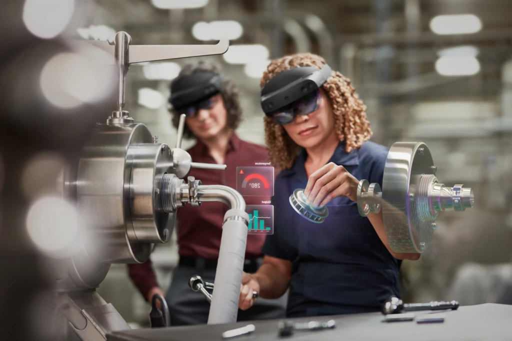 A woman wearing the HoloLens 2 fixing a machine. Another woman wearing a HoloLens also stands behind her