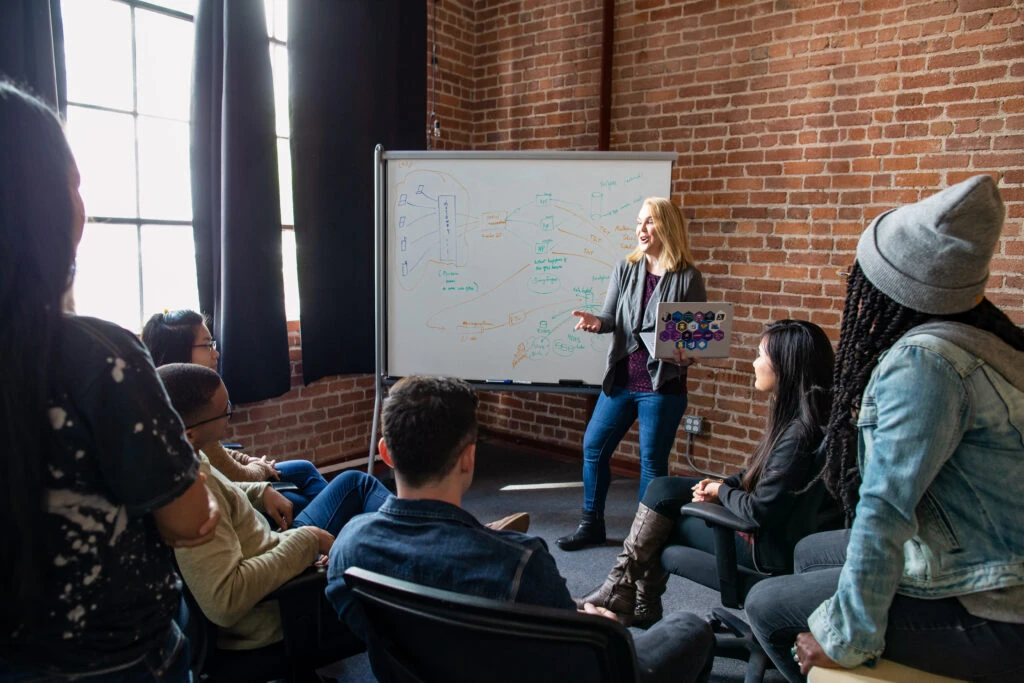 Female developer speaking in front of a white board during team stand up meeting, holding a Surface laptop personalized with stickers.