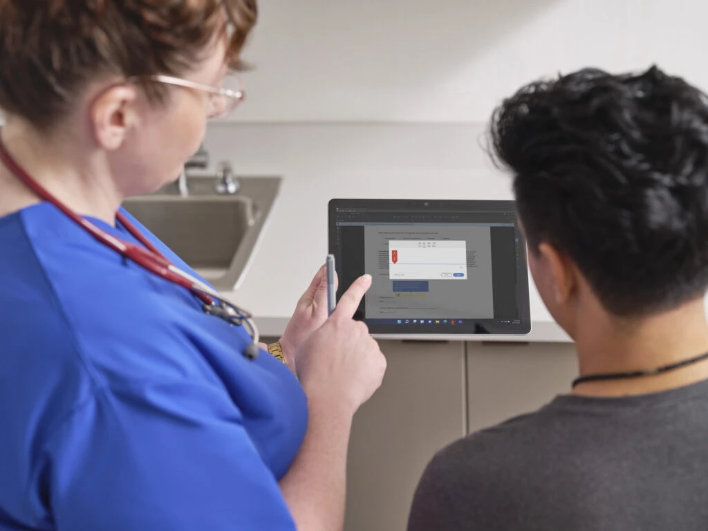 A female nurse is holding a Surface Go 3 and Surface Pen while instructing a patient where to sign. Screen shown is AdobeSign.