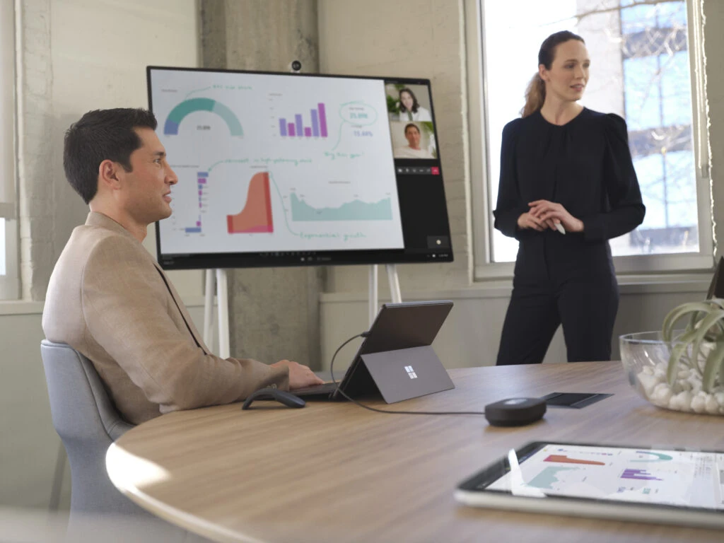 Two adults conducting a meeting in a conference room while collaborating over a Microsoft Teams video call presented on a Surface Hub 50” device. Two Surface Pro 8 devices are also shown. One in laptop mode with Surface Pro Signature Keyboard and one in tablet mode with Microsoft PowerBI shown.