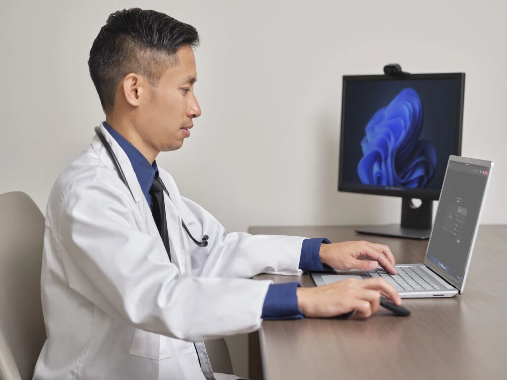 A healthcare professional is sitting at a desk using a Surface Laptop 4 with Teams on the screen. The monitor on the desk is displaying Window 11 screen.