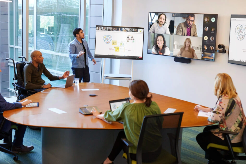 A coordinated meeting is taking place in a Microsoft Teams Room; people joining from the room and several joining remotely in Gallery view. A man is working on a Whiteboard; remote attendees can see the Whiteboard and collaborate. Two men and one woman joined the Teams meeting from their laptops and are able to write on the Whiteboard without having to move from the conference room table. 