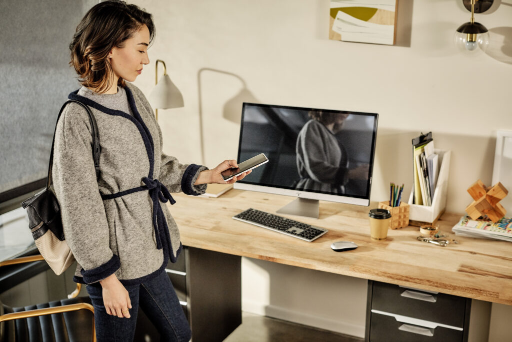 A woman checks her phone as she prepares to leave her office. A Acer desktop computer is in the background on her desk.