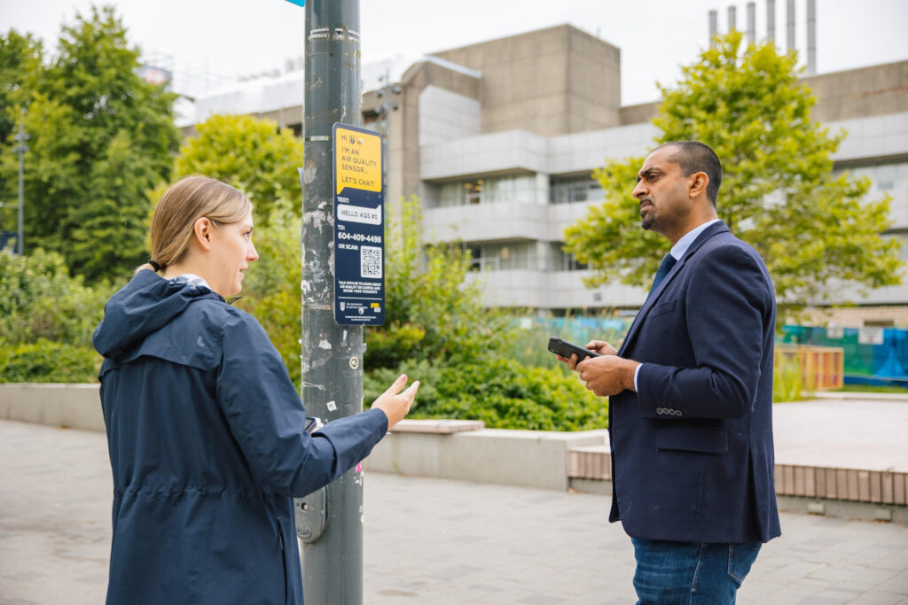 Two people using their phones stand next to a metal pole with a Hello Lamp Post sign on it, which states that it's an air quality sensor and they can chat with it.