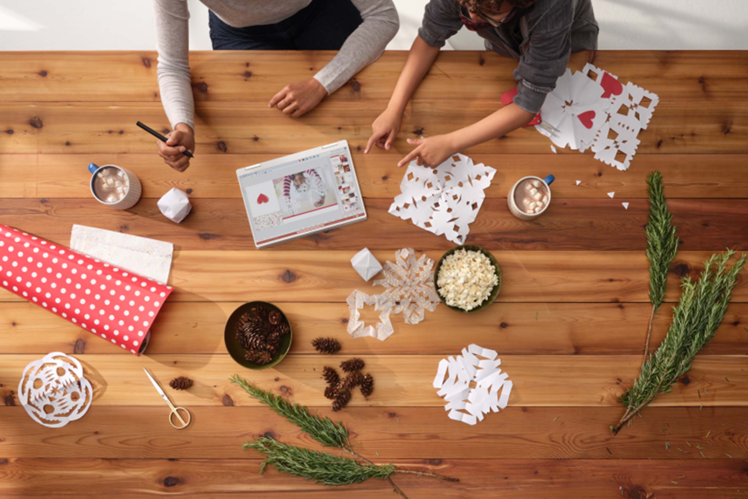 View from above of two people making paper snowflakes on a large wooden table.