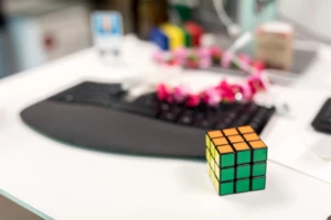 Sharp focus close-up of Rubik‘s cube on white desk. Black keyboard, pink flower lei, and other blurred items visible in background.
