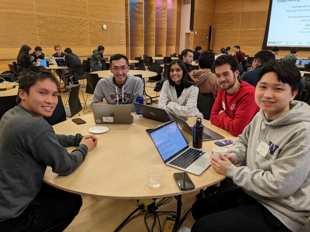 Five college age students sit at a table with their laptops, smiling at the camera as they participate in the NQN Hackathon.