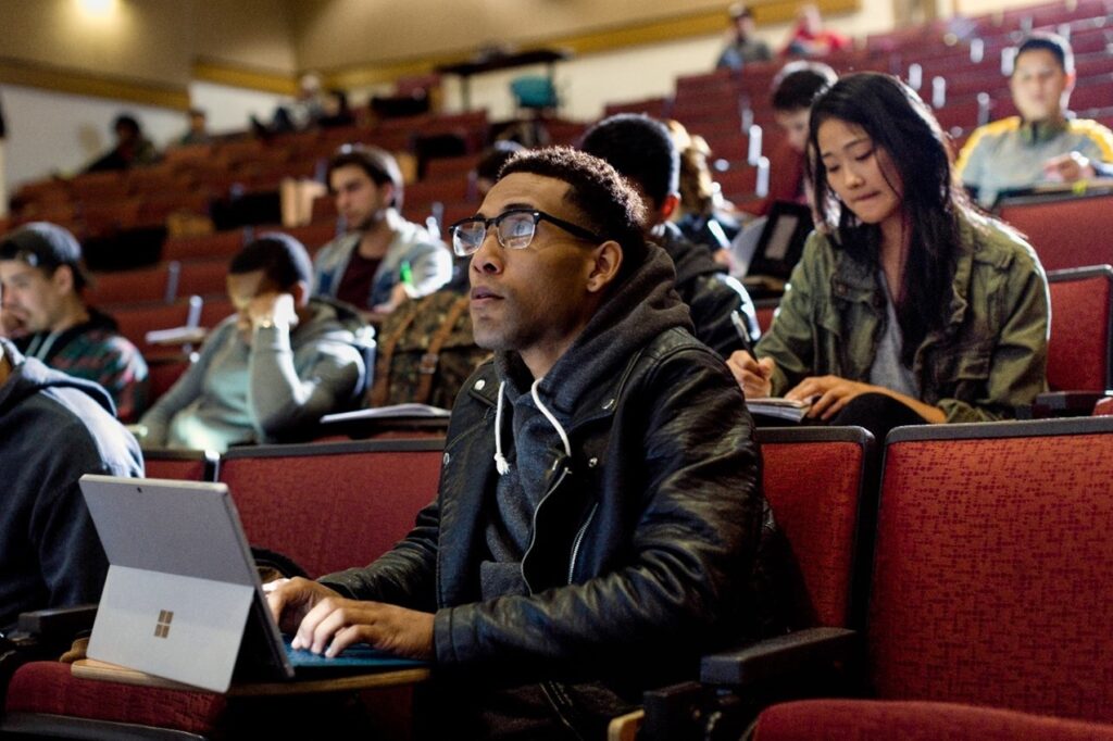 Close up of male college student sitting in university lecture hall, looking ahead while taking notes on a Surface Pro. Other male and female students sit behind him, taking notes.